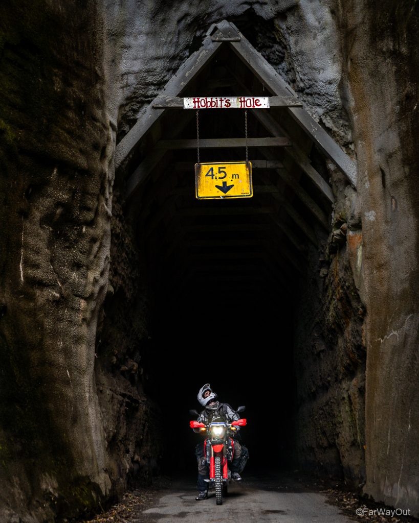Image of motorcyclist on red bike looking up at the'Hobbits Hole' sign as he comes out of Moki Tunnel along Forgotten World Highway on North Island of New Zealand